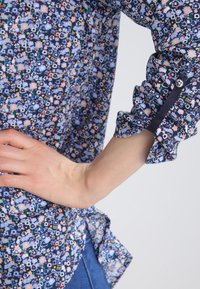 Arm bent with hand on hip, wearing a navy blue floral shirt with rolled-up sleeves secured by a buttoned tab.