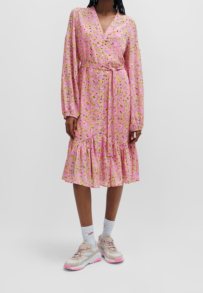 Femme portant une robe midi rose à manches longues à motif floral avec un ourlet volanté, taille nouée, chaussettes blanches et baskets beige-rose, debout devant un fond uni.
