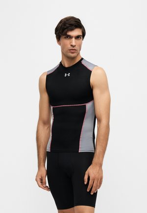 Jeune homme aux cheveux foncés portant un t-shirt de compression sportif sans manches noir et gris et un short noir ajusté, debout devant un fond uni.