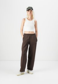 Hvit, cropped tanktop, brune joggebukser med lommer og hvite joggesko. Modellen har på seg en mørkegrønn caps med tekstur.