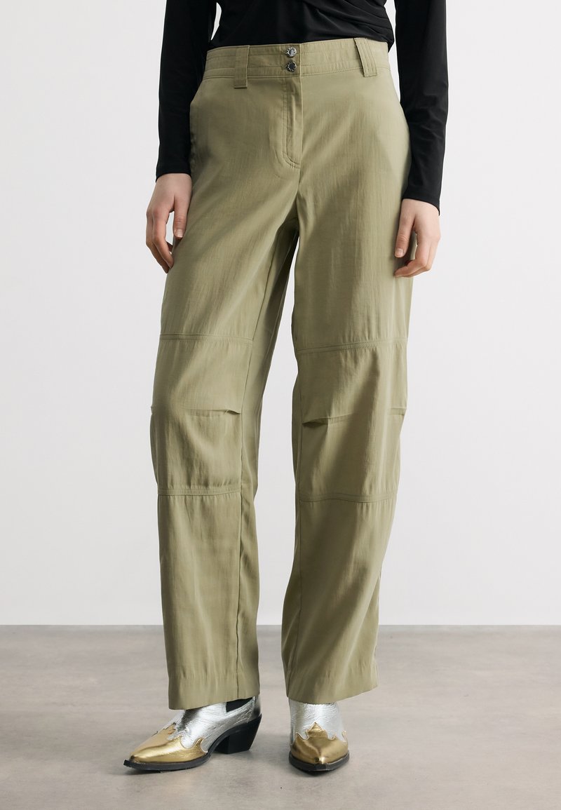 Γυναίκα που φοράει χακί παντελόνια με φαρδιά μπατζάκια, σε συνδυασμό με μεταλλικές ασημί και χρυσές μπότες αστραγάλου με μυτερή μύτη, όρθια σε απλό δάπεδο.