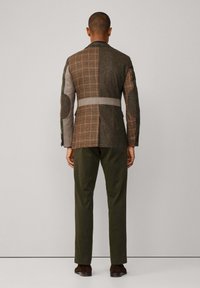 Multi-patroon blazer met bruine en grijze textielen, voorzien van een ruitpatroon, opvallende schouderpatches en een tailleband. Gecombineerd met groene broek.