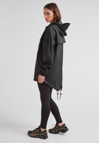 Svart vattentät jacka med dragskohuva, långa ärmar, sidofickor och asymmetrisk fåll, tillsammans med svarta leggings och sneakers.