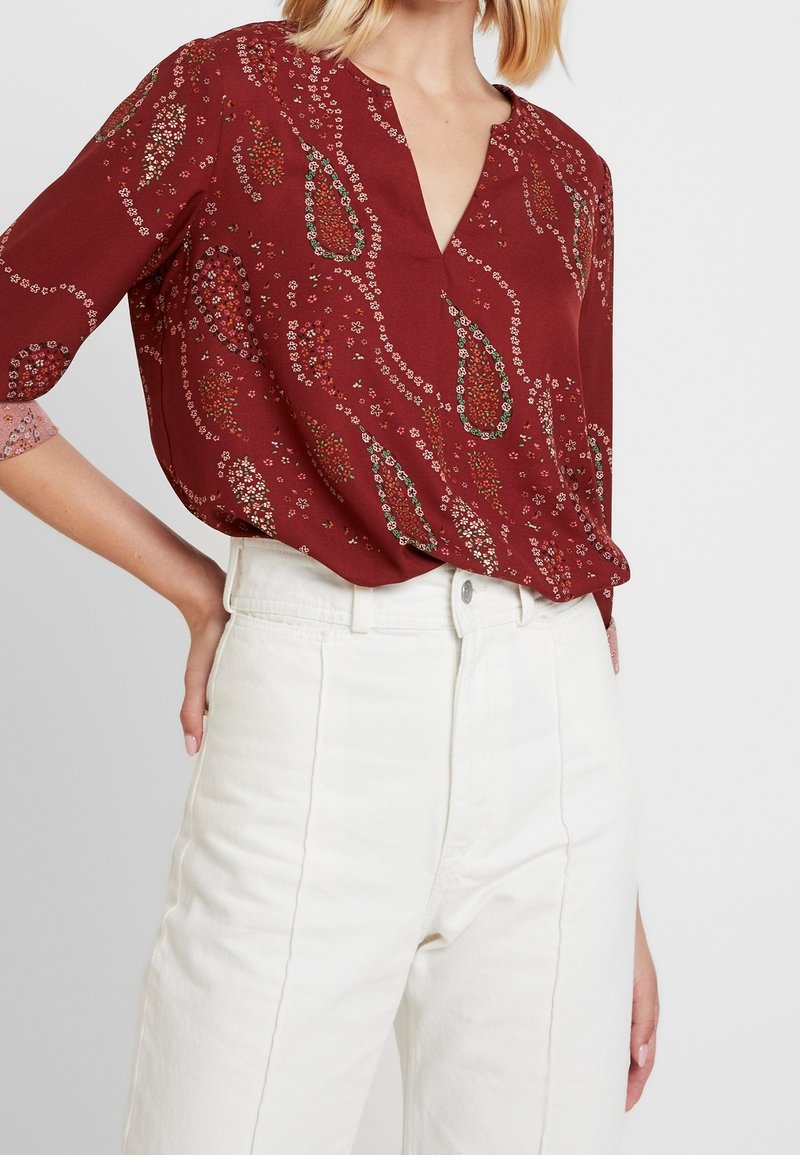 Blouse bordeaux avec un décolleté en V, des manches trois-quarts et un motif floral en paisley. Associée à un pantalon blanc taille haute. Texture lisse.