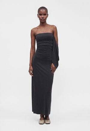 Femme portant une longue robe noire bustier avec une manche drapée et des chaussures plates beiges, debout devant un fond clair uni.