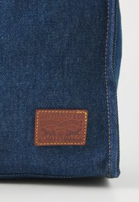 Denim tas in diepblauw met een gestructureerd oppervlak. Bevat een rechthoekige bruine leren patch met opdruk en gele stiksels.