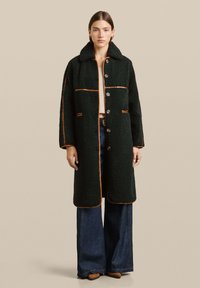 Donna con un lungo cappotto in pelliccia nera con rifiniture marroni, jeans blu scuro a gamba larga, maglione beige e scarpe a punta marroni, in piedi su uno sfondo beige.
