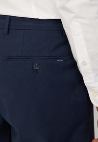 Pantalons navy en tissu texturé, dotés d'une poche arrière avec fermeture par bouton et du nom de la marque "Hackett" sur le côté.
