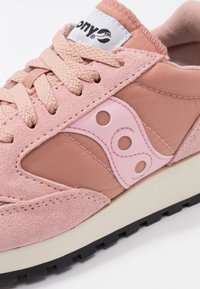 Chaussure de sport rose et beige avec panneaux en daim et tissu, lacets roses, languette avec logo, et semelle en caoutchouc noire avec motif de crampons.