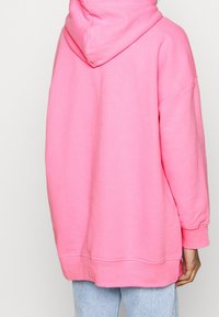 Rosa oversized hoodie med dragsko i huvan, tillverkad av mjukt tyg. Har breda ärmar och rundad nederkant. Bärs med ljusblå jeans.