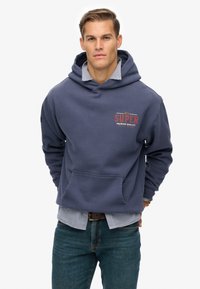 Man in een marineblauwe hoodie met de tekst 'dry SUPER premium quality', gelaagd over een geruit overhemd en gecombineerd met blauwe jeans, met de handen achter zijn rug.
