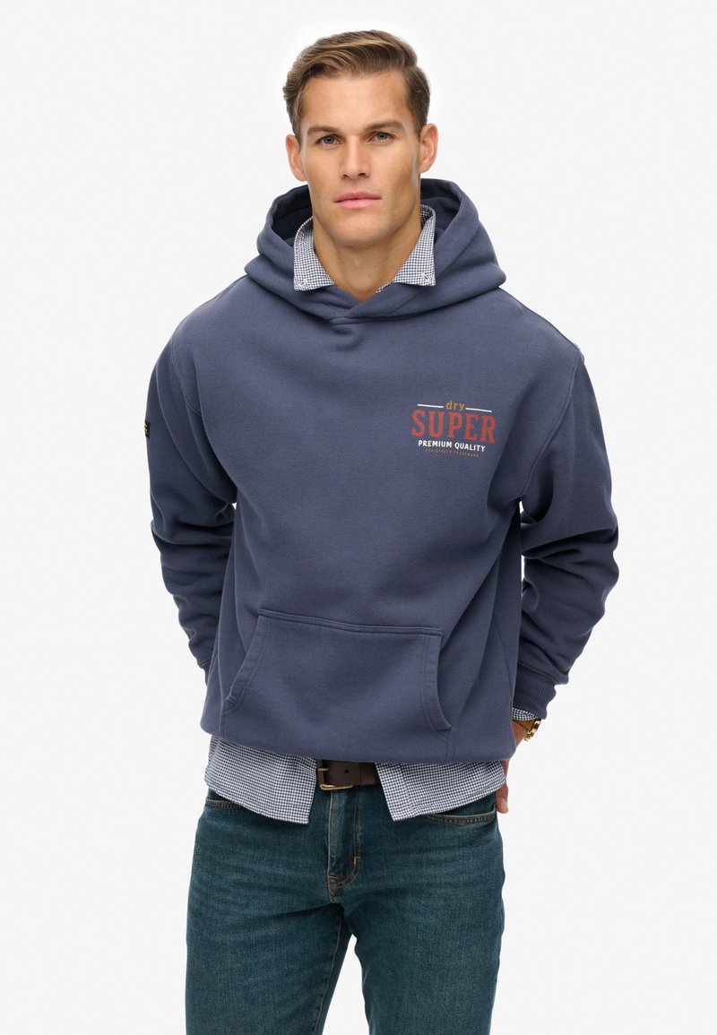 Man in een marineblauwe hoodie met de tekst 'dry SUPER premium quality', gelaagd over een geruit overhemd en gecombineerd met blauwe jeans, met de handen achter zijn rug.