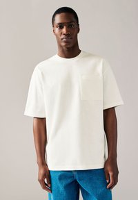 Kurzärmeliges, hellcremefarbenes T-Shirt mit Rundhalsausschnitt und Brusttasche, kombiniert mit blauen Jeans aus Denim. Flache, glatte Stoffstruktur.