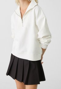Pull-over blanc avec col à zip, doté de poignets et d'un ourlet côtelés, associé à une jupe plissée noire, créant un contraste de couleurs.