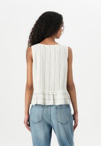 Bílý bezramínkový top s vertikálními béžovými pruhy, s peplum lemováním a uvolněným střihem. Kombinovaný se světlemodrými džíny s vysokým pasem.