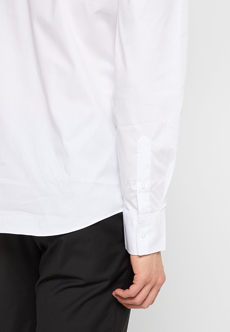 Chemise blanche à manches longues en tissu lisse, avec des poignets boutonnés et un ourlet droit. Portée avec un pantalon sombre, mettant en avant un design de poignet détaillé.