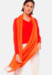 Écharpe orange avec des bords frangés, drapée sur un haut rouge. L'écharpe a une texture douce et est conçue pour être superposée. Un pantalon blanc complète le look.