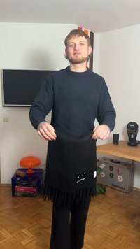 Zwarte sjaal met franjeranden en een logolabel, gemaakt van zachte stof. Model draagt een marineblauwe trui en zwarte broek, staand binnen.