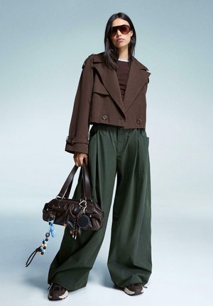 Giacca corta marrone con doppi bottoni, pantaloni a gamba larga verde scuro, borsa oversize marrone con charm colorati e sneakers marroni.