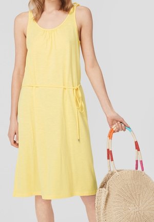 Frau trägt ärmelloses gelbes Kleid mit Taillenbindung und hält eine runde, gewebte Tasche mit mehrfarbigen, gestreiften Griffen.