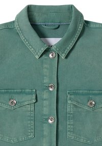 Veste en denim vert vue de face avec des boutons-pression argentés et deux poches poitrine avec rabats, montrant le col et la partie supérieure du vêtement.