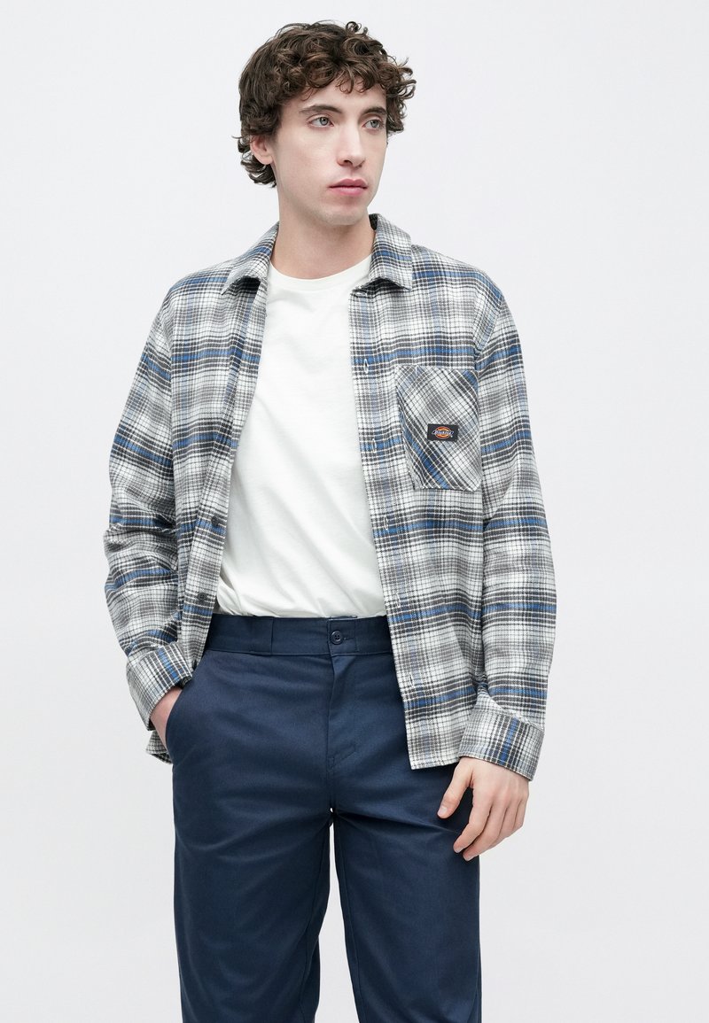 Graues und blau kariertes Hemd mit Brusttasche, getragen über einem weißen T-Shirt, kombiniert mit marineblauen Hosen. Glatter Stoff, Knopfleiste.