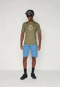 Olīvu zaļš īsās piedurknes krekls ar logo, apvienots ar gaiši ziliem šortiem. Aksesuāri ietver melnu ķiveri un saulesbrilles, kā arī melnas sporta kurpes.