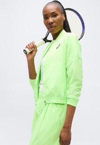 Γυναίκα με νέον πράσινη φόρμα, κρατώντας ρακέτα του τένις ακουμπισμένη στον ώμο της, στέκεται μπροστά σε λευκό φόντο.