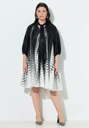 Donna in piedi che indossa un vestito al ginocchio con motivo bianco e nero, maniche a tre quarti e décolleté abbinate con punta appuntita.