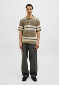 Maglietta a maniche corte lavorata a maglia di colore marrone con strisce bianche e beige; presenta un colletto a punta e bottoni; indossata con pantaloni larghi scuri e scarpe nere.