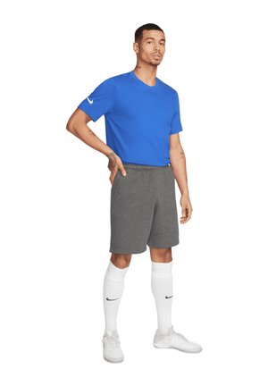 Blauw korte mouwen sportshirt, grijze shorts, witte kniekousen met logo en witte sportschoenen. Model staat met een ontspannen houding.