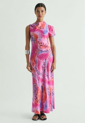 Mujer con vestido sin mangas hasta el suelo con estampado rosa, naranja y púrpura, sandalias negras y brazalete ancho plateado.