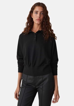 Zwart lange mouwen polo trui met een lage zoom en placket met drie knopen; textiel stof; verstelbare manchetten; gedragen met zwarte broeken.
