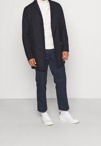 Manteau long bleu marine avec boutons, col roulé blanc, pantalon bleu marine et baskets blanches. Le manteau présente une coupe droite et un col cranté.