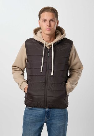 Jeune homme les mains dans les poches, portant un sweat à capuche beige et un gilet matelassé noir, debout devant un fond clair uni.