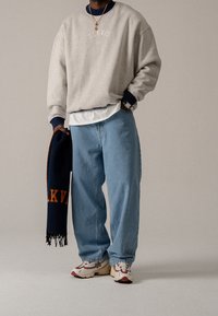 Světlě šedá mikina s tmavě modrými manžetami, modré džíny a vícebarevné tenisky. Drží tmavě modrý šátek s oranžovým nápisem.