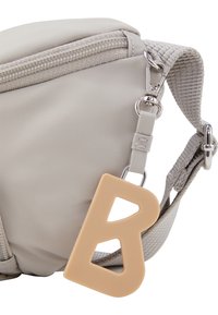 Harmaa nailonlaukku vetoketjulla, jossa on teksturoidulla pinnalla ja ruskean silikonisen "B"-koristeen, joka on kiinnitetty metalliklipsillä.