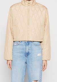 Beżowa krótka kurtka puchowa z pikowaniem w kształcie rombów, wysokim kołnierzem, zamkiem błyskawicznym i ściągaczami na rękawach. W zestawieniu z postrzępionymi niebieskimi jeansami.