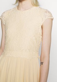 Kvinna iklädd en ärmlös beige klänning med blommig spetsliv och plisserad kjol, visar en tatuering i svart skrivstil på sin underarm.