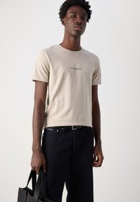 Tricou din bumbac bej cu text imprimat negru "Arrivederci", asortat cu blugi negri și ținând o geantă de umăr neagră.