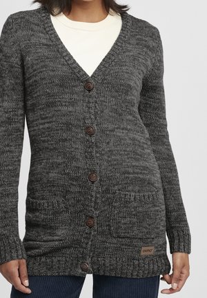 Cardigan en maille gris foncé à boutons, avec deux poches devant, porté sur un haut blanc et associé à un pantalon en velours côtelé bleu.