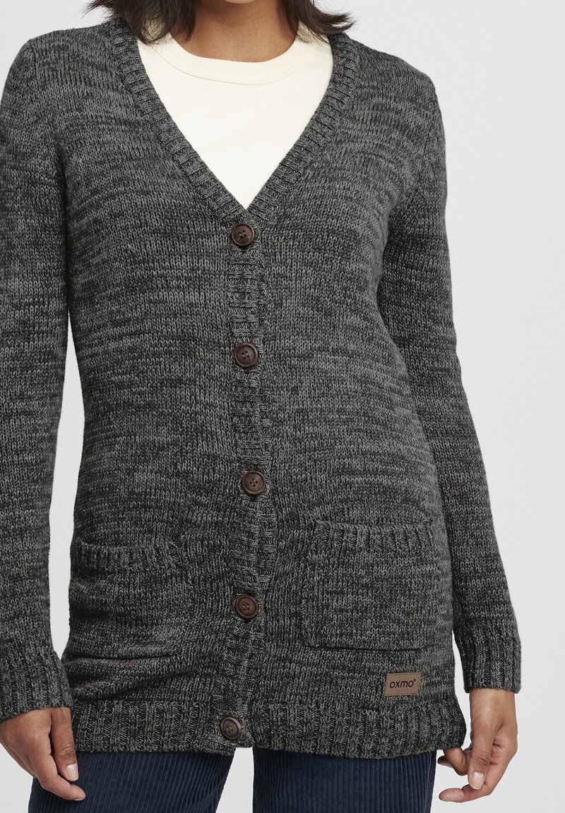 Cardigan en maille gris foncé à boutons, avec deux poches devant, porté sur un haut blanc et associé à un pantalon en velours côtelé bleu.