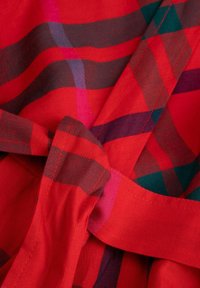 Zbliżenie czerwonej tkaniny z wzorem w kratę w kolorach czarnym, różowym i ciemnoniebieskim, pokazujące szczegółowe szwy i zawiązany supeł.