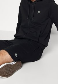 Felpa nera con zip e pantaloni corti abbinati in un tessuto morbido, con un piccolo logo di coccodrillo verde sul petto e sull'orlo. Sneakers bianche con suole testurizzate.