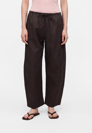 Personne portant un pantalon ample marron foncé à cordon, un haut rose clair et des sandales noires, debout devant un fond blanc uni.