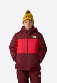 Red and maroon insulated jacket with a hood, featuring a zip front and two side pockets. Yellow patterned knit beanie with a pom-pom on top.