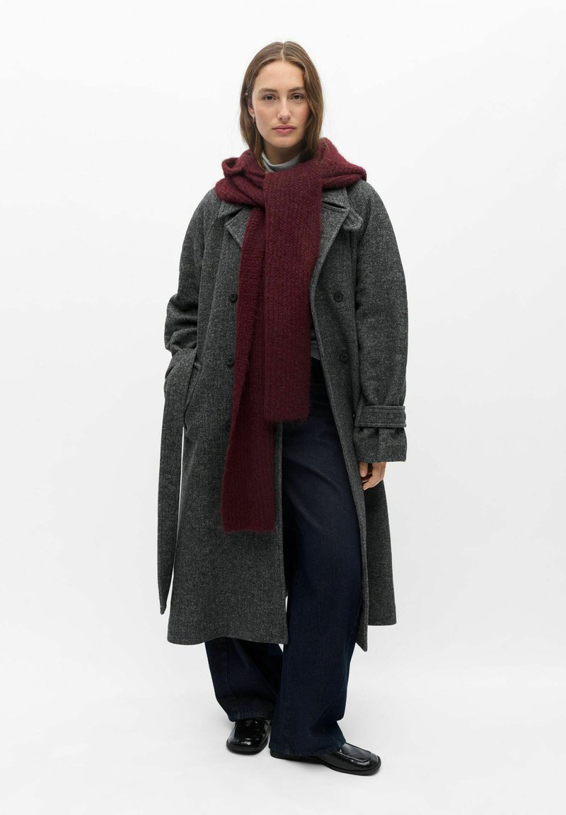 Femme debout portant un long manteau gris, un pantalon large foncé, des chaussures noires et une grande écharpe rouge foncé enroulée autour du cou.