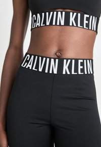 Milieu de la personne portant un soutien-gorge de sport noir Calvin Klein et des leggings avec de larges ceintures de marque, montrant la peau nue et le nombril.
