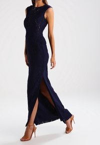 Robe en dentelle bleu marine avec une silhouette moulante, des manches courtes, des fentes latérales et un motif texturé, associée à des talons ouverts couleur nude.