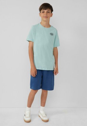 Lichtblauw katoenen T-shirt met een klein gedrukt logo, gecombineerd met donkerblauwe shorts en witte sneakers met beige accenten.
